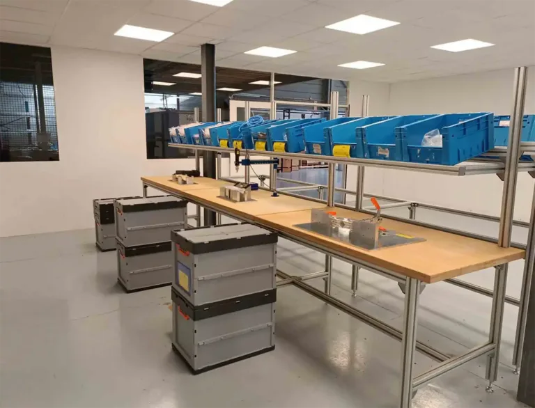 Industrial workspace with a long wooden workbench, blue plastic bins on an overhead rack, and gray tool chests nearby in a clean room.