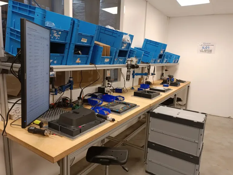 Electronics workbench with blue plastic storage bins, a tall monitor, and assorted tools along a long laboratory-style table.
